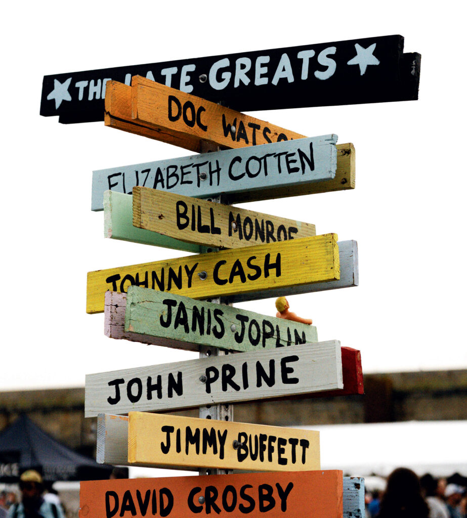 Newport Folk Festival in Rhode Island. A colorful wooden signpost with arrows naming musicians including Doc Watson, Johnny Cash, Janis Joplin, and others, under a sign reading "The Late Greats.