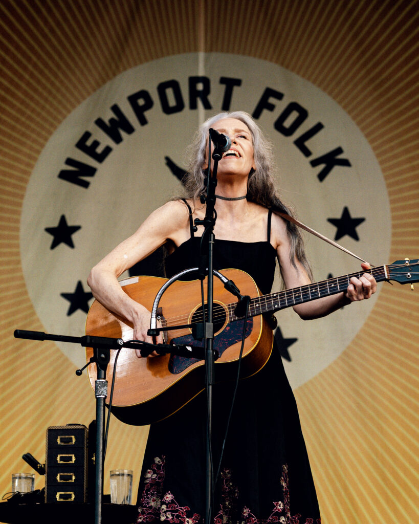 A woman with long gray hair plays an acoustic guitar and sings on stage in front of a "Newport Folk" festival sign.