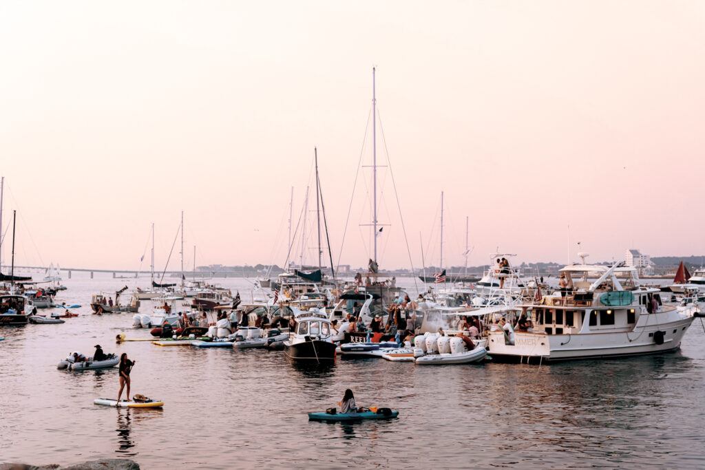 Several boats and yachts are anchored close together in a harbor at sunset, with people gathered on the vessels and a few individuals on paddleboards in the water.