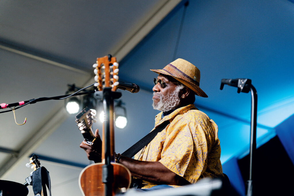 Newport Folk Festival in Rhode Island. An older man wearing a straw hat and yellow shirt plays an acoustic guitar and sings into a microphone on a stage under a tent.