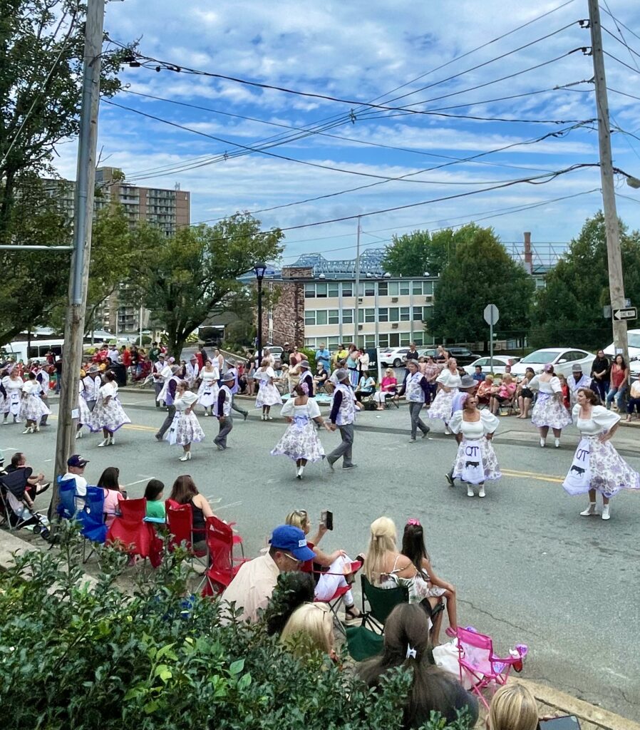 A group of people in matching outfits dance in formation on a street during a parade, while spectators watch from chairs and sidewalks.