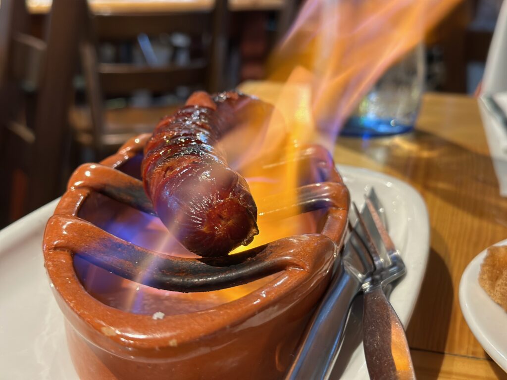 Grilled sausage served on a ceramic dish with open flames, accompanied by a fork and knife on a white plate.