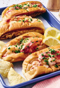 Warm Lobster Rolls with Sherry Butter. Three lobster rolls topped with chopped chives are served in a blue tray with potato chips and lemon wedges.