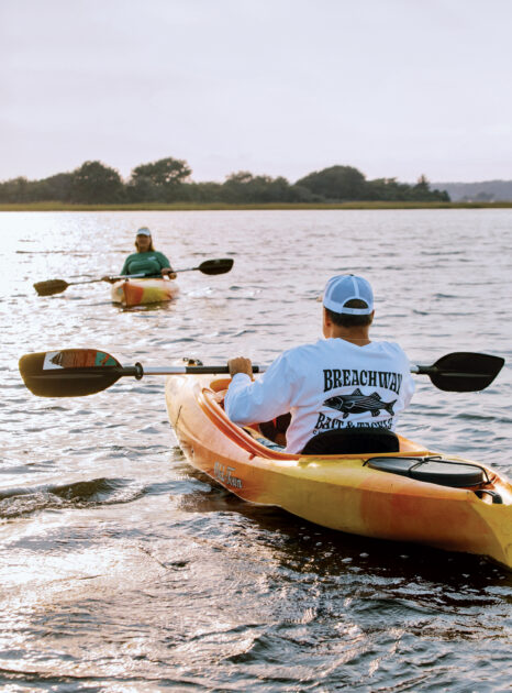 Two people are kayaking on a calm body of water during daylight, with trees visible in the distance.