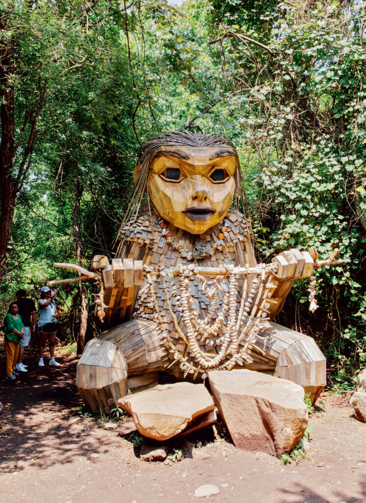 Things to Do in Charlestown, RI: Where to Eat, Stay & Play by the Beach. Large wooden sculpture of a troll-like figure sits in a forest clearing, adorned with necklaces and surrounded by trees, with people standing nearby for scale.