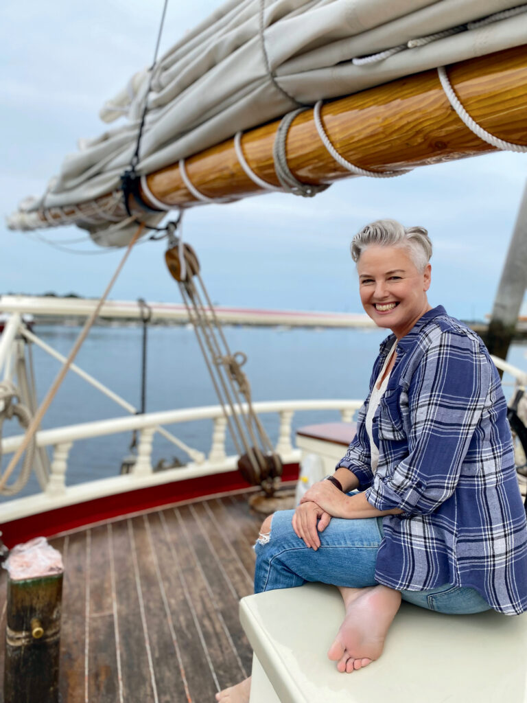 A smiling person with short gray hair and a plaid shirt sits barefoot on a bench aboard a sailboat, with rigging and calm water visible in the background.