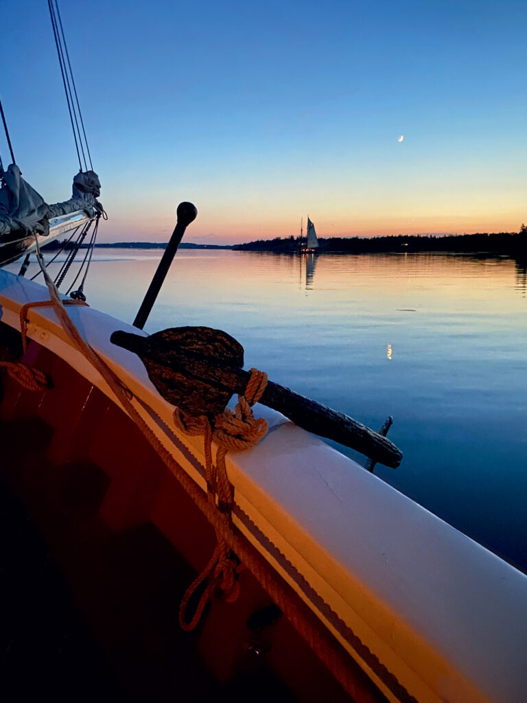 Sailing Memories on a Maine Windjammer. View from a boat at sunset, showing a wooden oar tied to the railing, calm water, a distant sailboat, and a crescent moon in the sky.