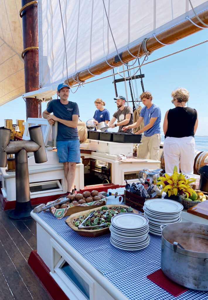 Sailing Memories on a Maine Windjammer. A group of people prepare and serve food on the deck of a sailing ship, with dishes and stacked plates set out on a table in the foreground.