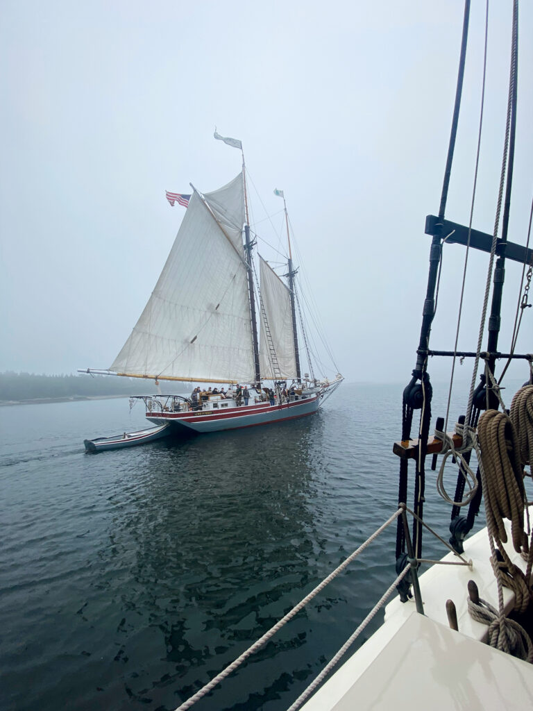 A large sailboat with an American flag sails on calm water in foggy weather, viewed from the deck of another boat.
