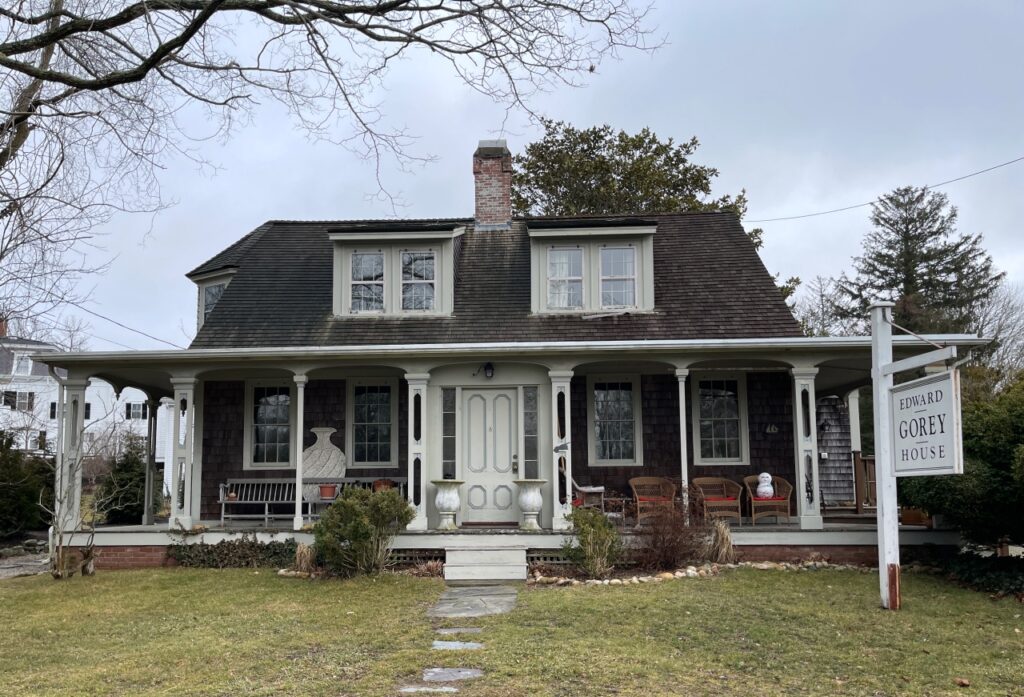 Edward Gorey House. A two-story historic house with a covered front porch, wooden chairs, and a sign reading "Edward Gorey House" on a cloudy day.