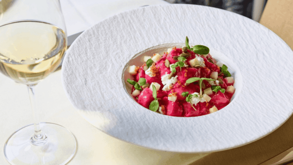 A glass of white wine next to a bowl of vibrant pink beet gnocchi topped with herbs and cheese.