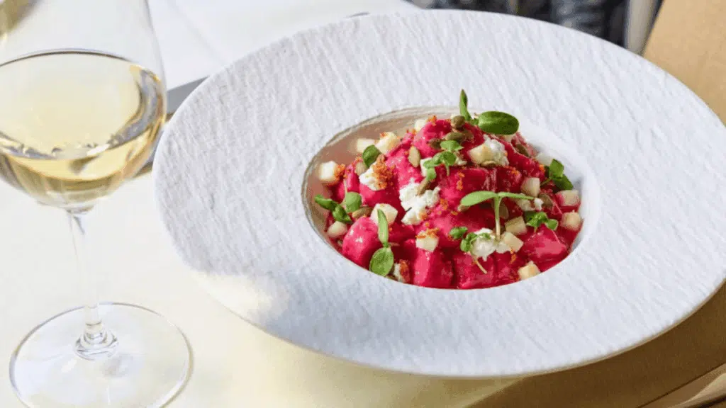 A glass of white wine next to a bowl of vibrant pink beet gnocchi topped with herbs and cheese.
