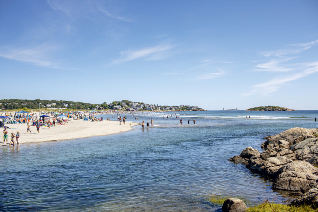 The Great Ocean vs. Lake Debate. People walk and relax on a crowded sandy beach next to calm ocean water on a sunny day, with rocks in the foreground and houses visible in the background.