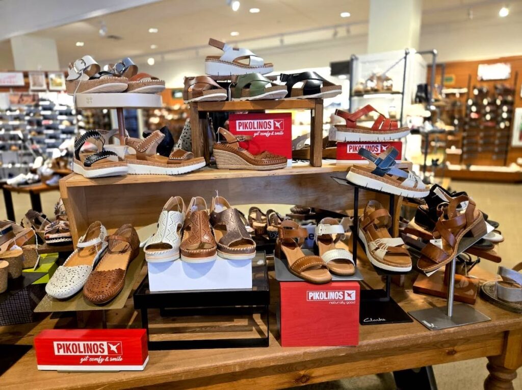 Display of various Pikolinos sandals in neutral tones arranged on wooden tables and shelves inside a well-lit shoe store.