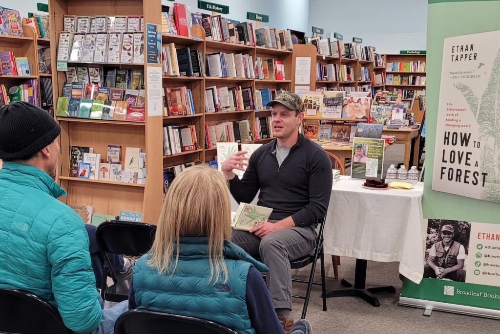 An author speaks to a small group at a bookstore event, holding a book titled "How to Love a Forest" with a promotional banner and display beside him.