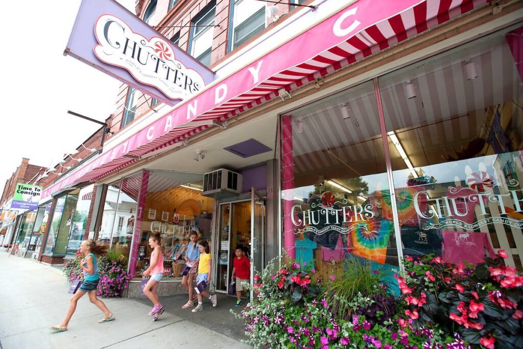 Children exit a colorful candy store called Chutters on a busy street, with vibrant awnings, flower planters, and passersby on the sidewalk.