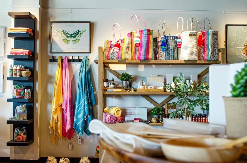 A shop display with colorful striped bags, towels, lotions, and self-care products on shelves and racks, with a framed butterfly print on the wall.