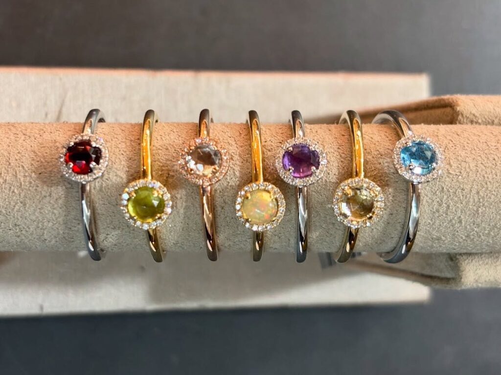Six gemstone rings with different colored stones and small clear accents are displayed on a beige ring holder against a dark background.