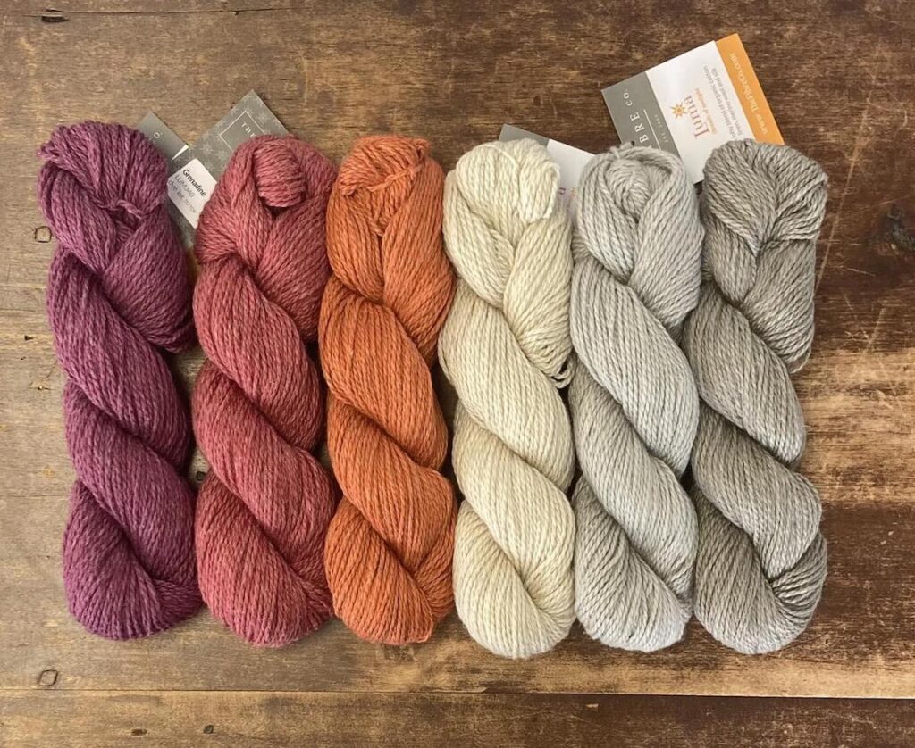 Six skeins of yarn in shades of purple, red, orange, cream, and gray are arranged side by side on a wooden surface.