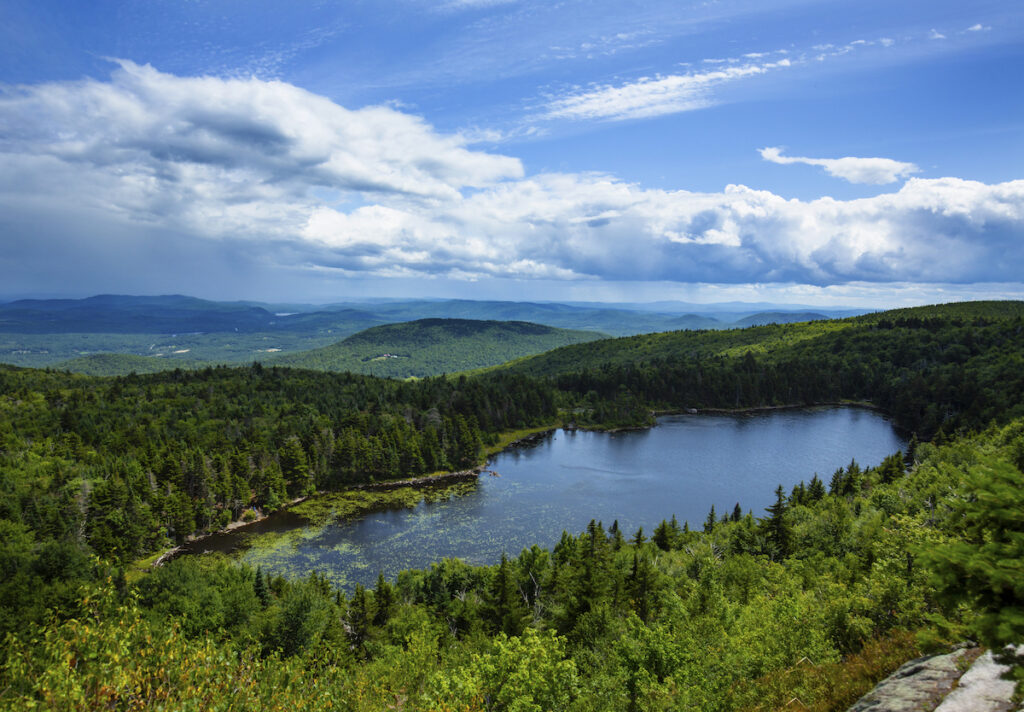 A small, tree-lined lake is surrounded by dense green forest with rolling hills and a partly cloudy sky in the background.