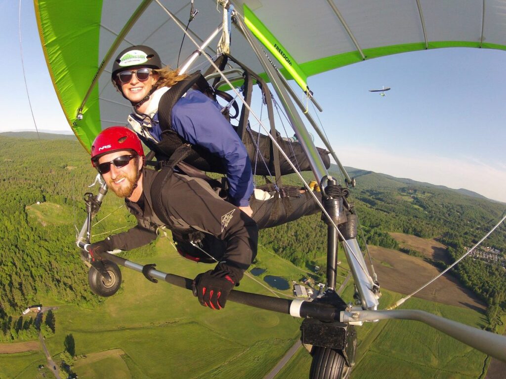 Two people are tandem hang gliding high above a green landscape, wearing helmets and sunglasses, with trees and fields visible below.