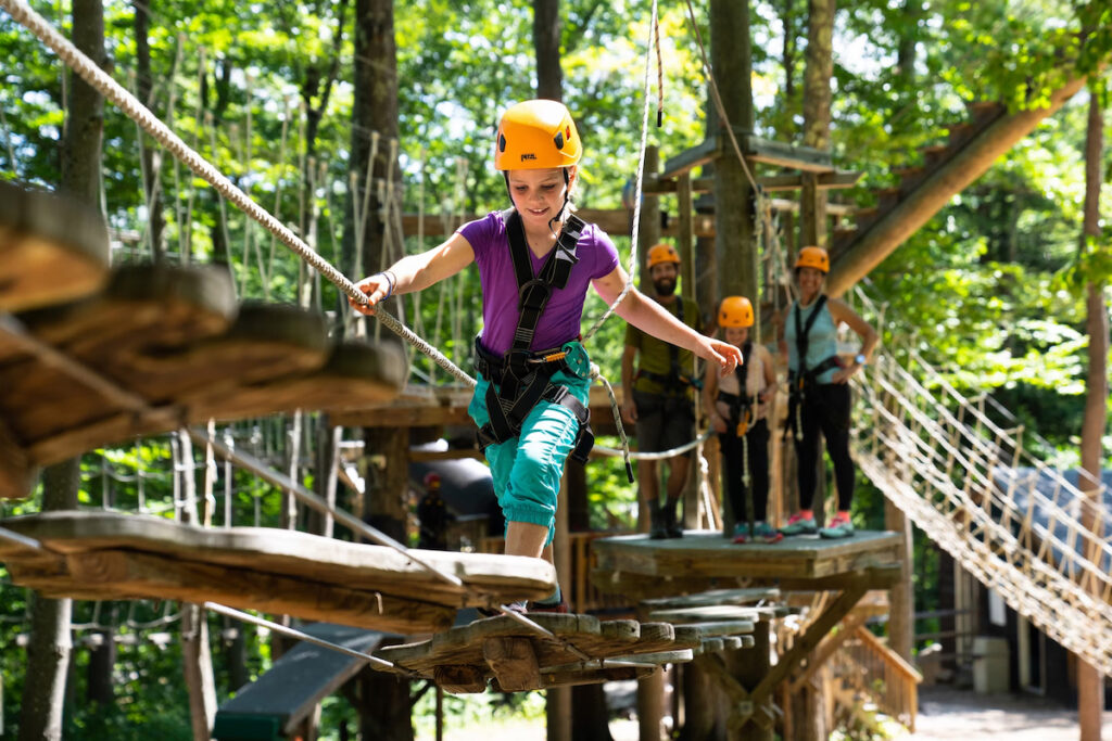 A child wearing a helmet and harness crosses a wooden bridge in a forest adventure park, while others wait behind on a rope bridge.