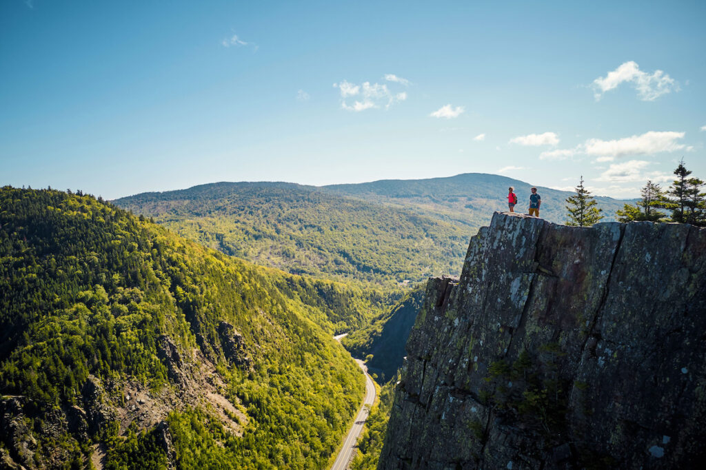 Two people stand on the edge of a cliff overlooking a forested valley and distant mountains under a clear blue sky.