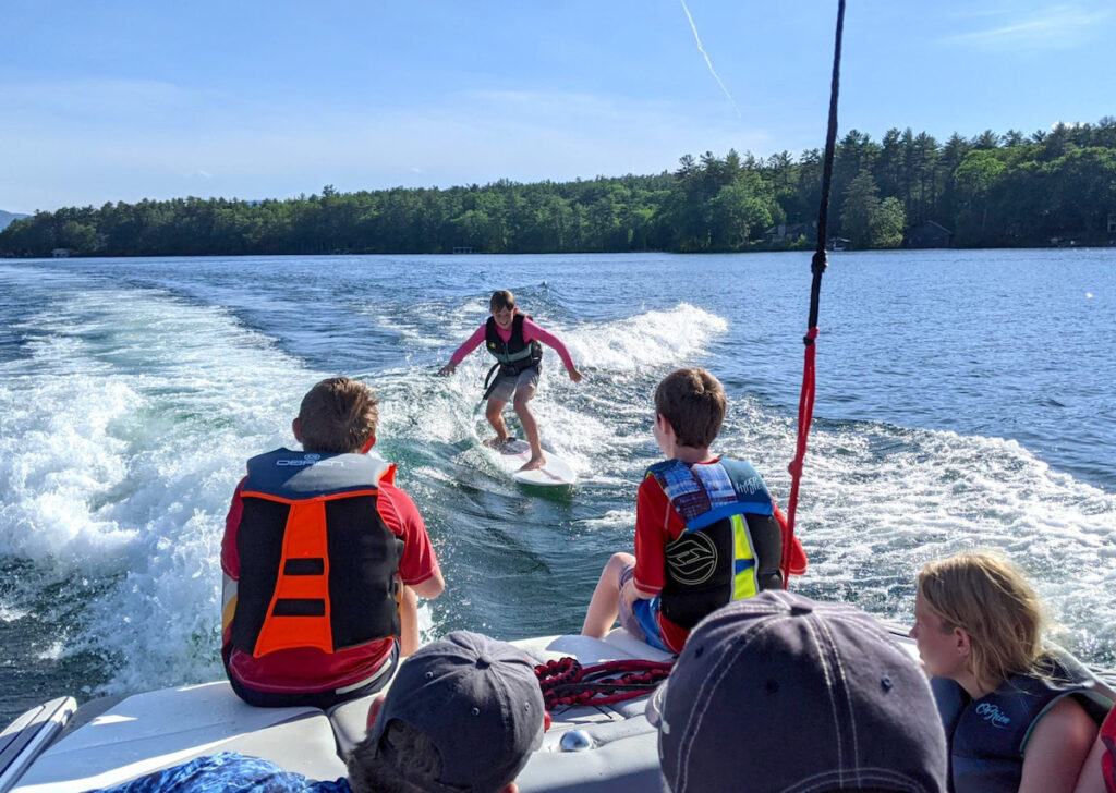 A person surfs behind a boat on a lake while three children in life jackets watch from the boat. Trees and a clear sky are visible in the background.