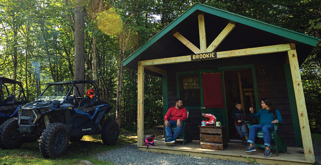 Three people sit on the porch of a small cabin labeled "Brookie," with two off-road vehicles parked nearby and trees in the background.