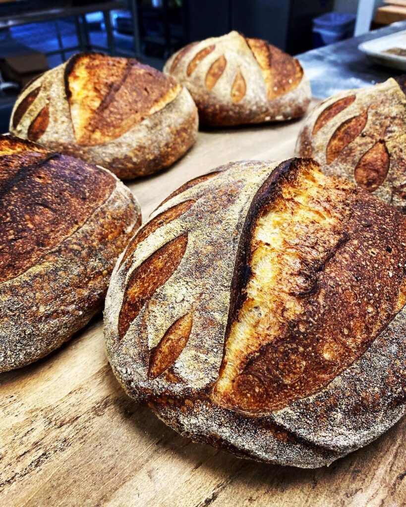 Several round loaves of rustic sourdough bread with dark, crusty tops are cooling on a wooden surface in a bakery setting.