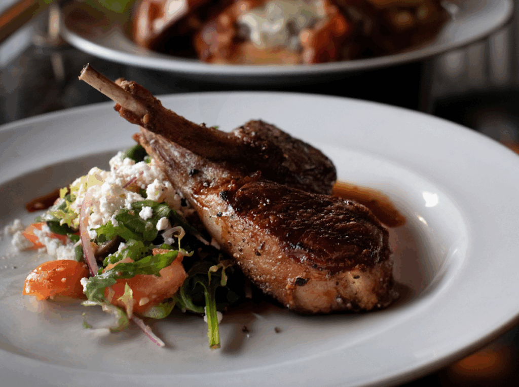 Two grilled lamb chops served on a white plate with a side salad of mixed greens, tomatoes, onions, and crumbled cheese.