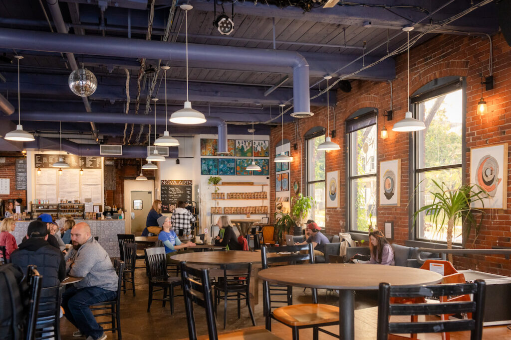 A spacious coffee shop with brick walls, large windows, people sitting at tables, and a counter in the back.