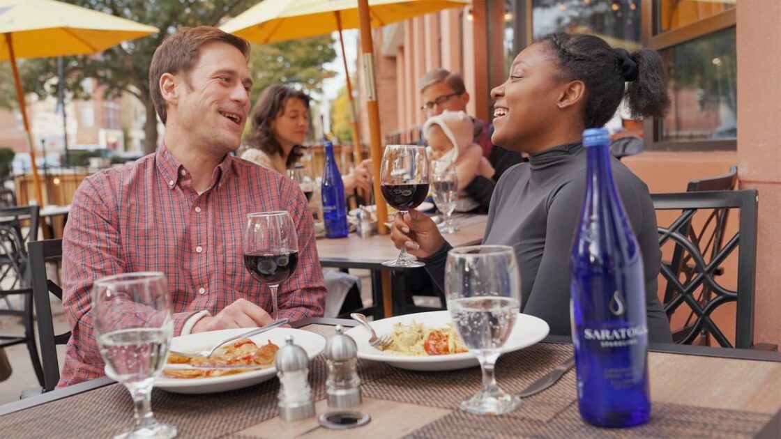 Two people sit at an outdoor restaurant table, smiling and holding wine glasses, with plates of food and a blue water bottle in front of them. Other diners are seated in the background.