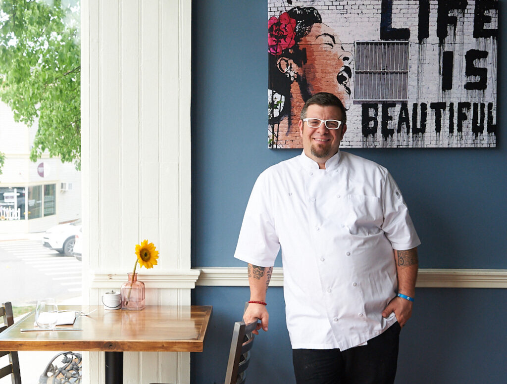 A chef in a white uniform stands by a table with a sunflower in a vase, next to a wall with a "Life is Beautiful" mural.