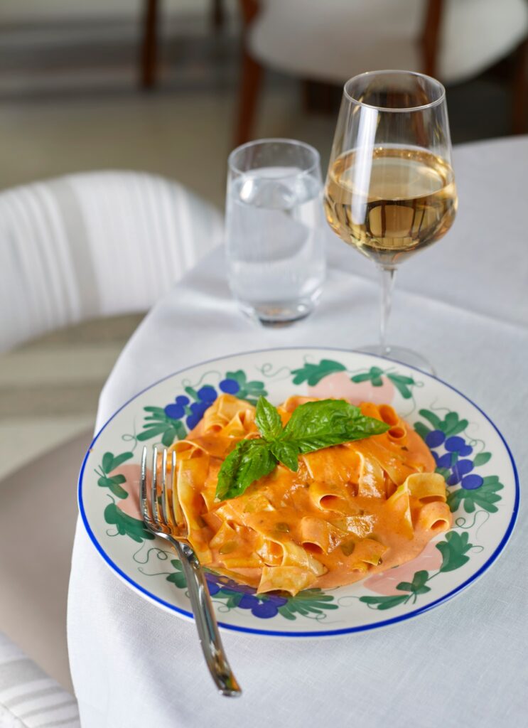 A plate of pasta in creamy tomato sauce garnished with basil, served with a fork, a glass of white wine, and a glass of water on a white tablecloth.