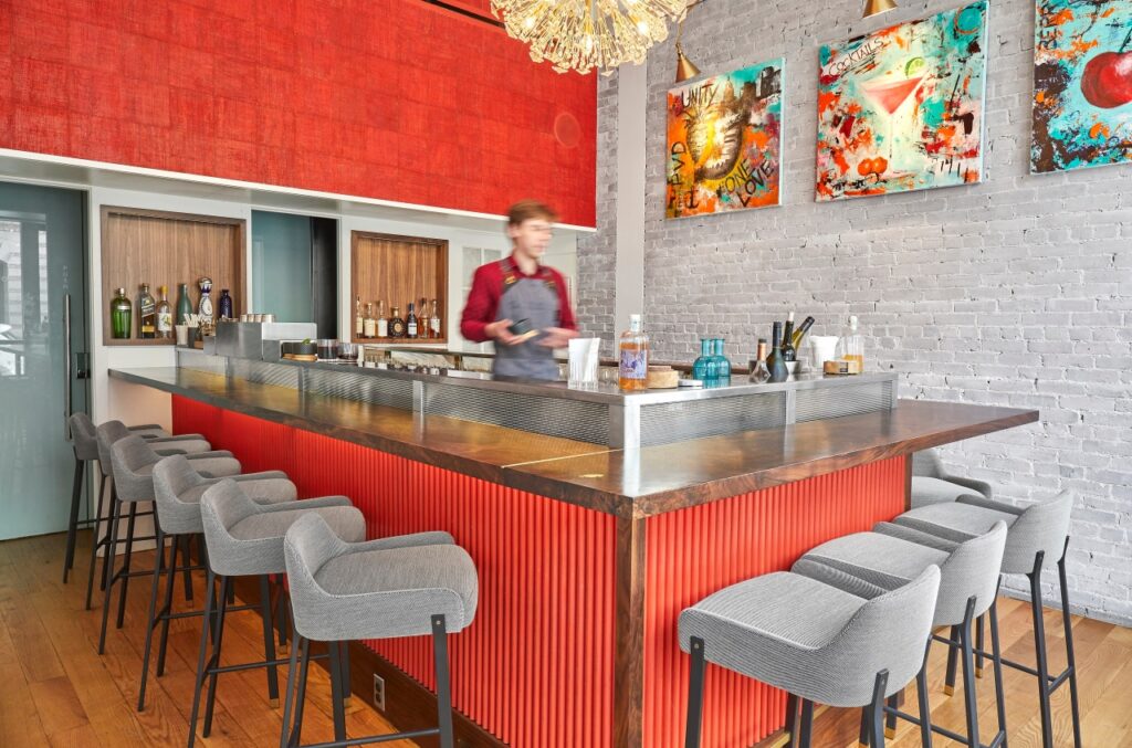Modern bar with red accents, gray chairs, and a bartender standing behind the counter; abstract art and bottles decorate the background.