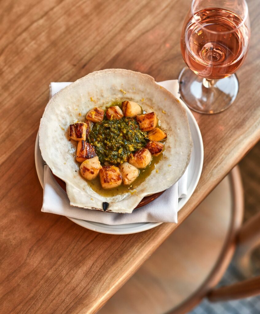 A plate with seared scallops arranged in a shell around a green sauce, served on a napkin with a glass of rosé wine on a wooden table.