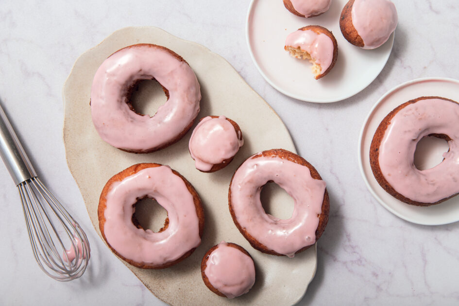 Maine Potato Doughnuts with Cranberry Glaze. Pink-glazed donuts and donut holes are arranged on a plate and a small dish, with a whisk covered in pink glaze set to the side on a white marble surface.