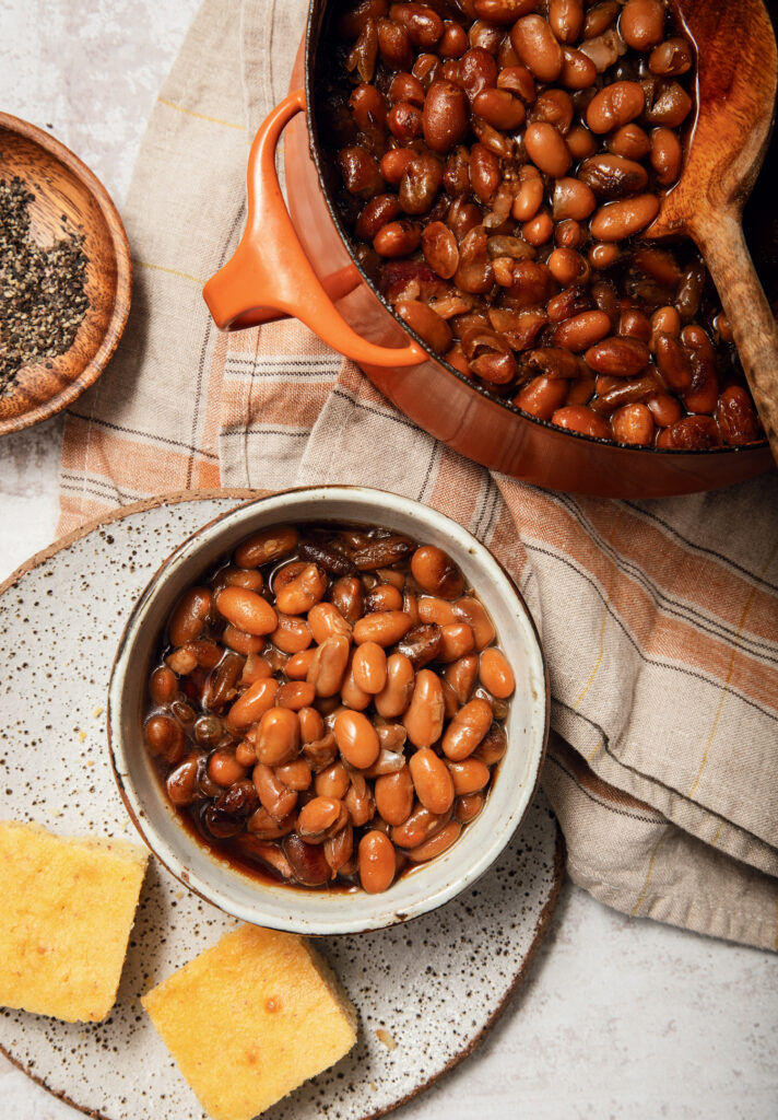 Durgin-Park Baked Beans. A bowl of baked beans in sauce on a speckled plate with two cornbread squares, next to a pot of beans and a small dish of black pepper.
