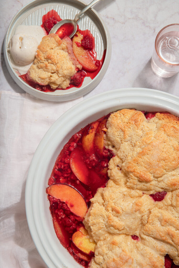 Louisa May Alcott’s Fruit Slump. A peach and raspberry cobbler in a white dish, with a serving of cobbler and a scoop of vanilla ice cream on a plate nearby.