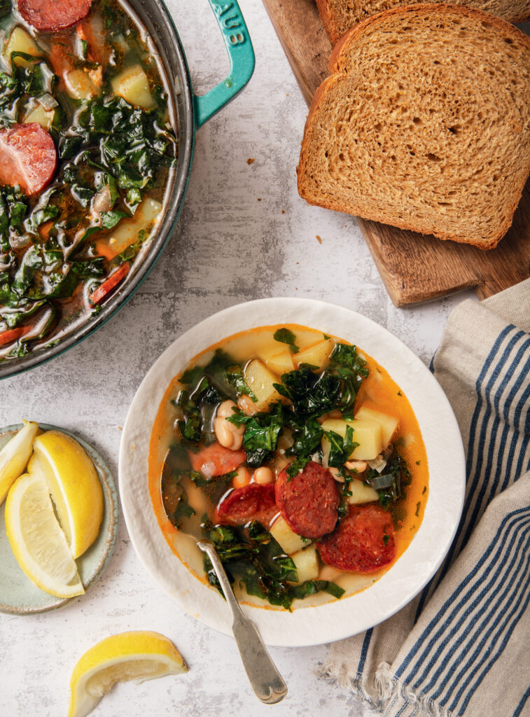 A bowl of soup with sausage, kale, beans, and potatoes sits next to slices of whole wheat bread and lemon wedges on a textured surface.