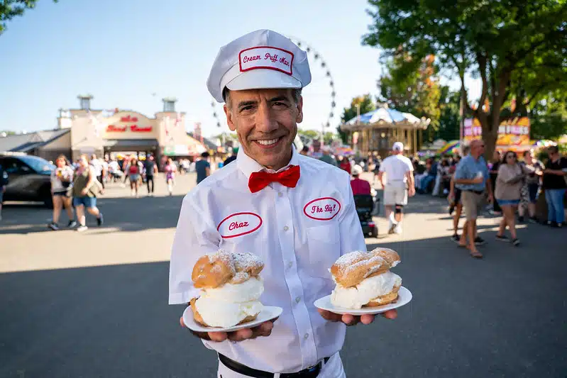 Smiling man in a white uniform holds two plates of cream puffs at The Big E.