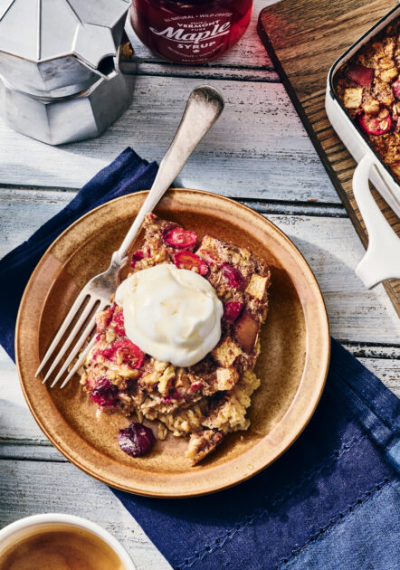 A plate with a serving of baked oatmeal topped with yogurt, a fork, and a pot of coffee, with maple syrup and a baking dish in the background.
