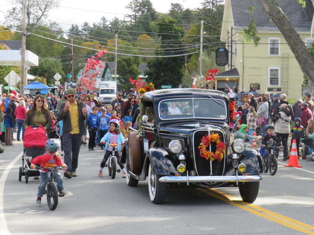 A vintage car decorated with autumn leaves drives in a parade, surrounded by children on bicycles and a crowd of spectators on a small town street.