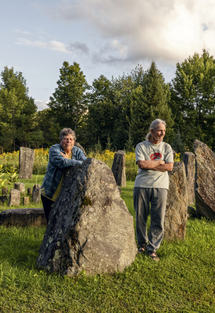 Two older adults stand outdoors among large upright stones on a grassy area, with trees and a partly cloudy sky in the background.