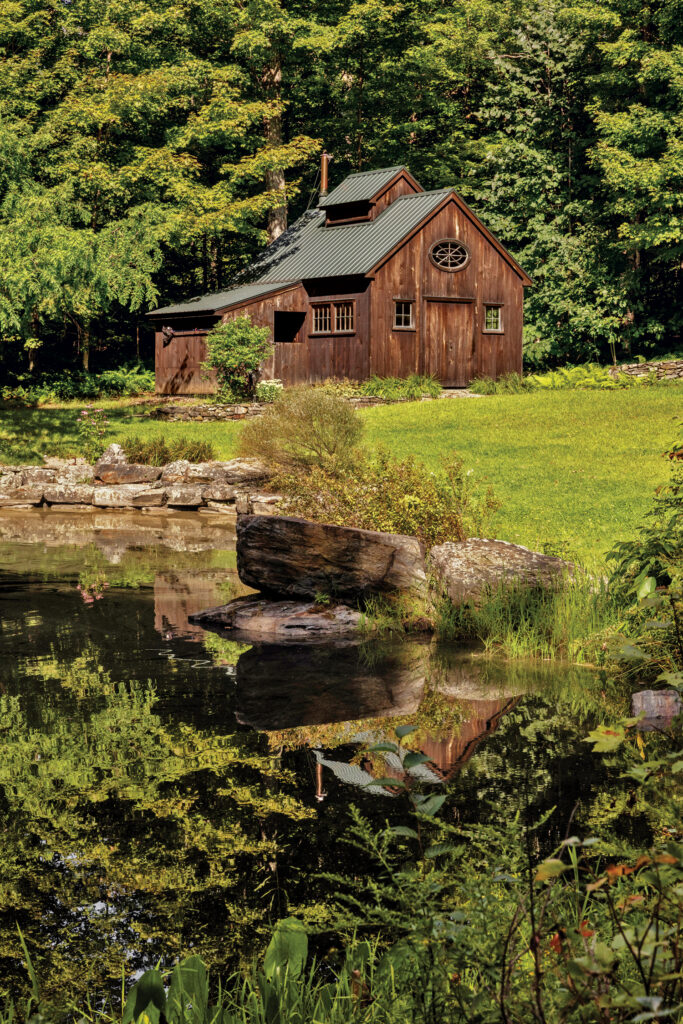 A rustic wooden cabin stands beside a pond, surrounded by green grass, rocks, and dense trees in the background.