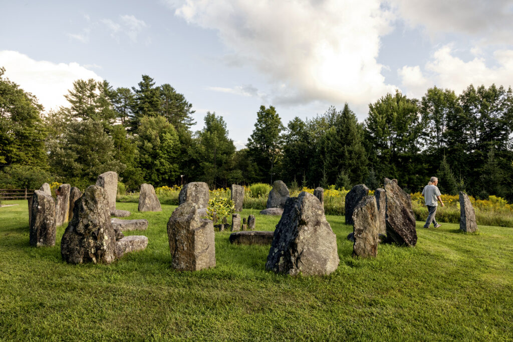 A circular arrangement of large upright stones on grass, surrounded by trees, with a person walking nearby under a partly cloudy sky.