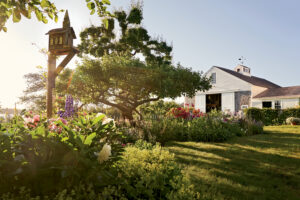 A lush garden with blooming flowers, a large tree, a wooden birdhouse, and a white barn-style building under clear blue sky with sunlight streaming through.