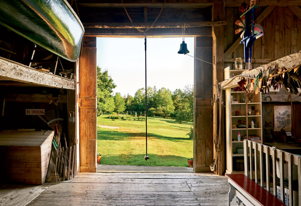 View from inside a rustic wooden barn looking out onto a grassy field, with a canoe overhead, shelves on the right, and a rope hanging from the ceiling.