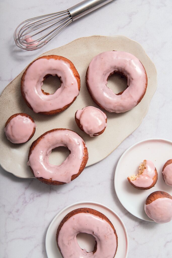 A plate and small dish with pink-glazed donuts and donut holes, one partially eaten, next to a whisk with pink glaze on a light marble surface.